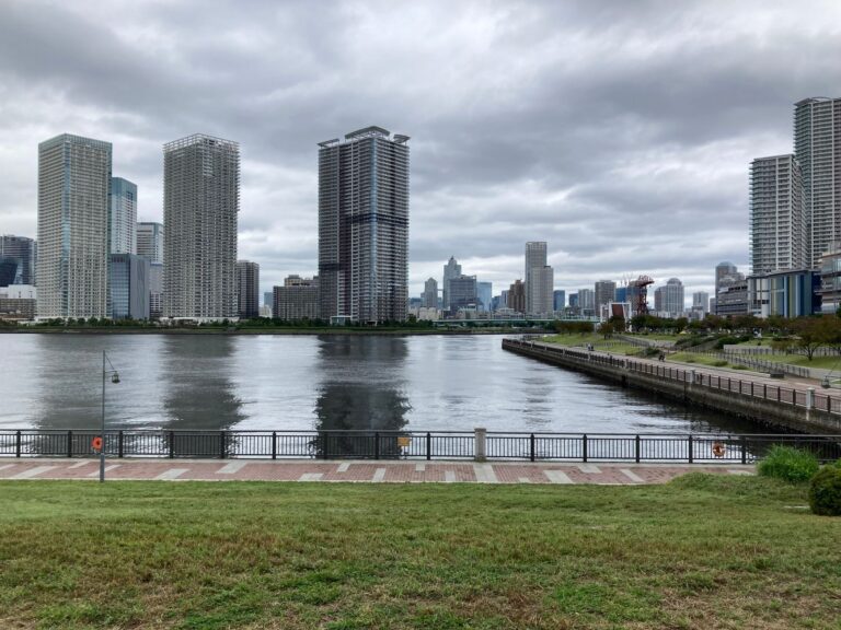 Scenic view of Tokyo skyline from Odaiba, featuring modern skyscrapers and waterfront promenade under cloudy skies.
