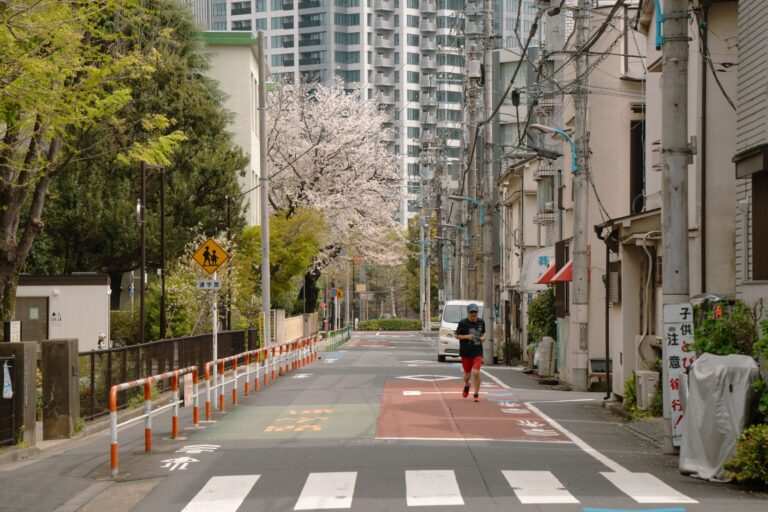 A person jogging on a tree-lined street in Tokyo with cherry blossoms and urban buildings in view.